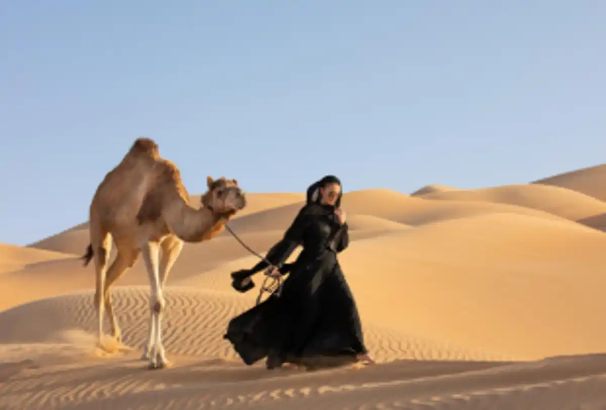 Woman walking with camel across golden desert dunes at sunset