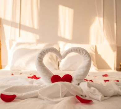 Romantic honeymoon bedroom setup with heart-shaped towel swans and red rose petals on white bed linens.